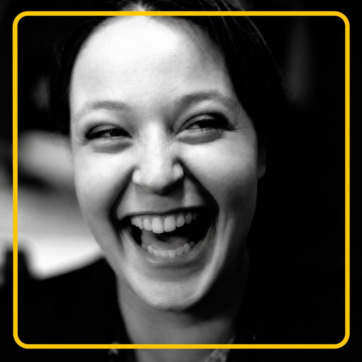 Portrait en noir et blanc d'une femme aux cheveux sombres qui rit de bon cœur, entouré d'un cadre jaune.